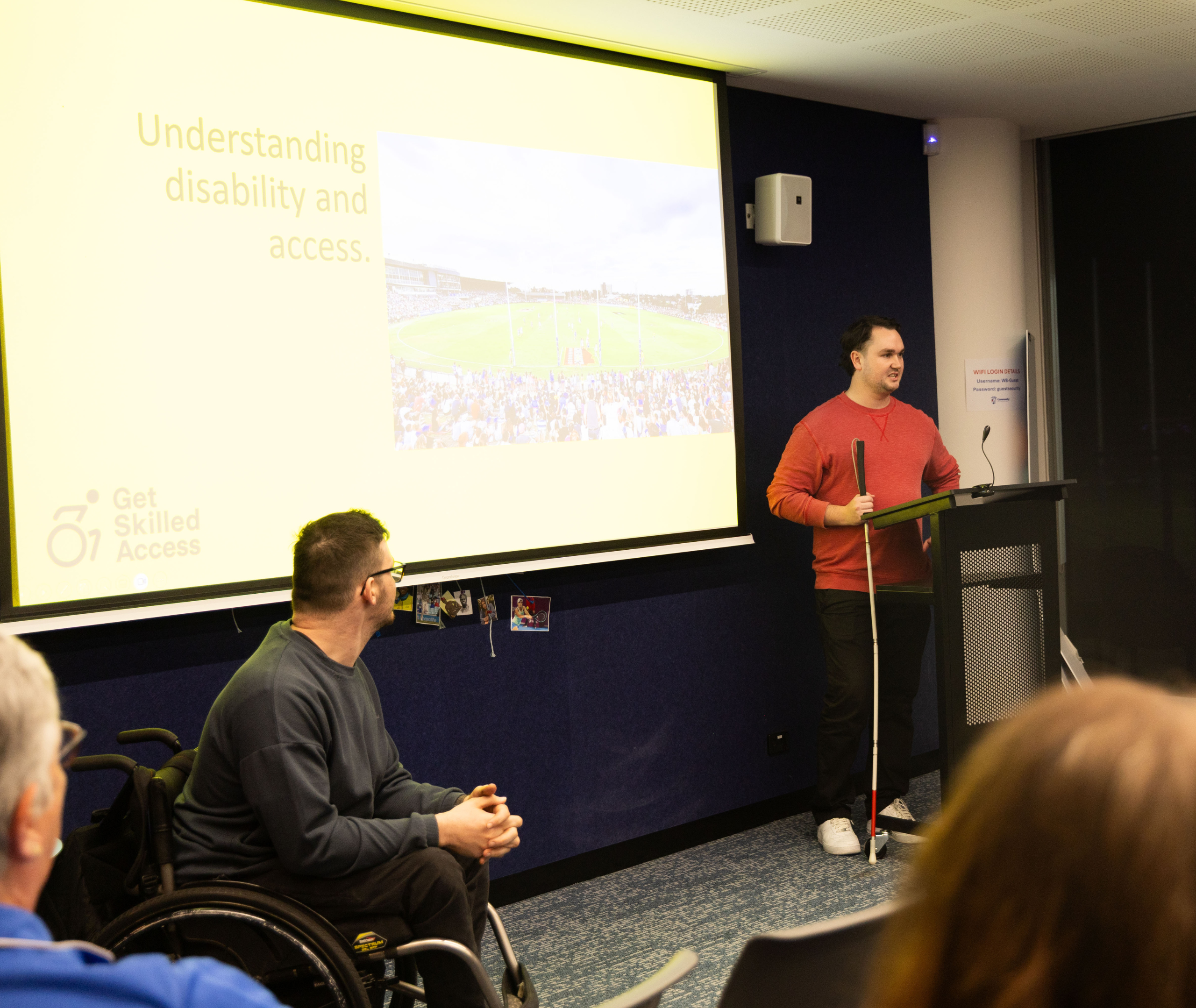 Johnny standing at a lectern with a white cane presenting a slide titled “Understanding disability and access” with Oliver looking seated on the wheelchair.