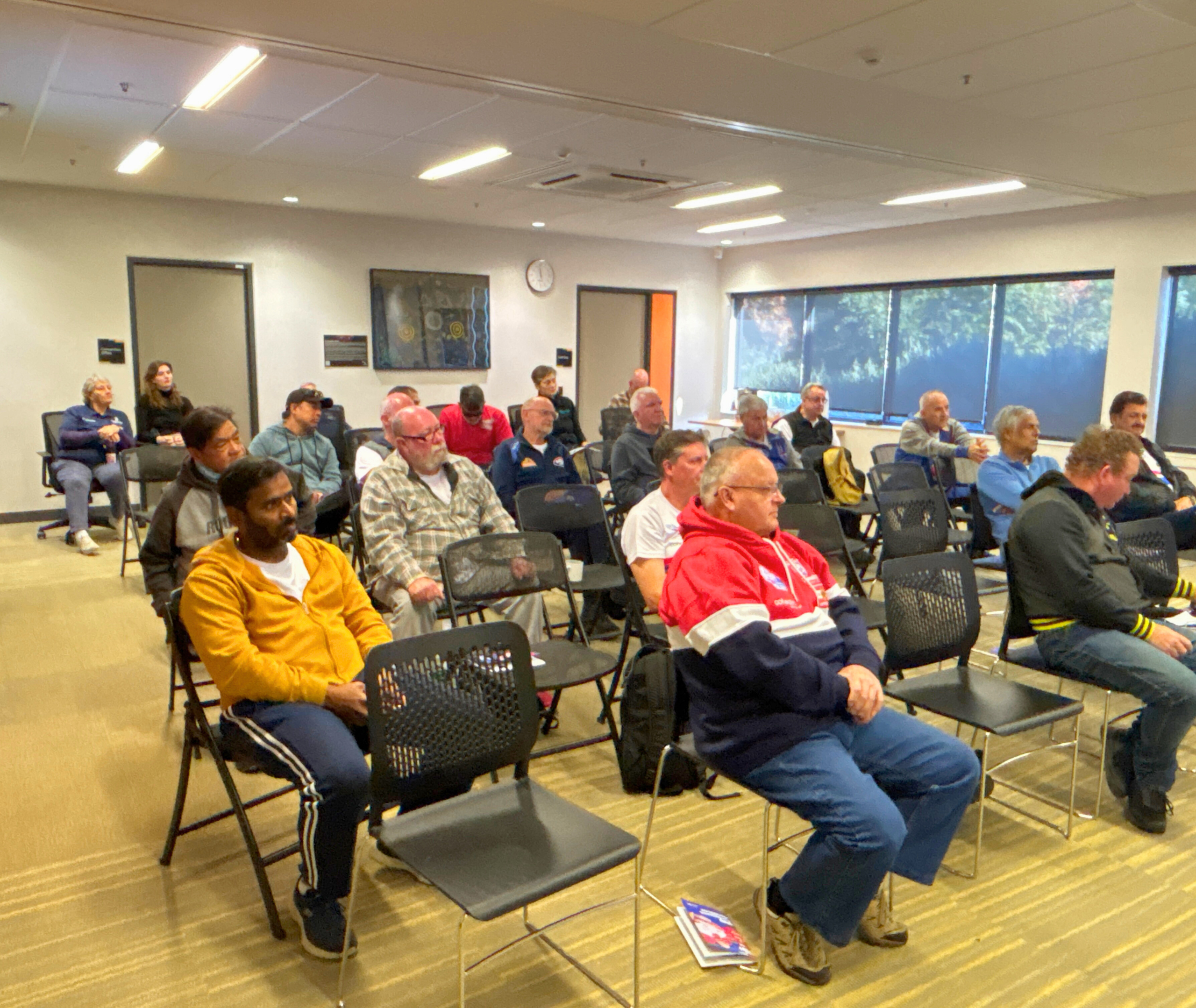 A mixed group of participants sitting in rows in a meeting room, listening attentively to a speaker out of frame.