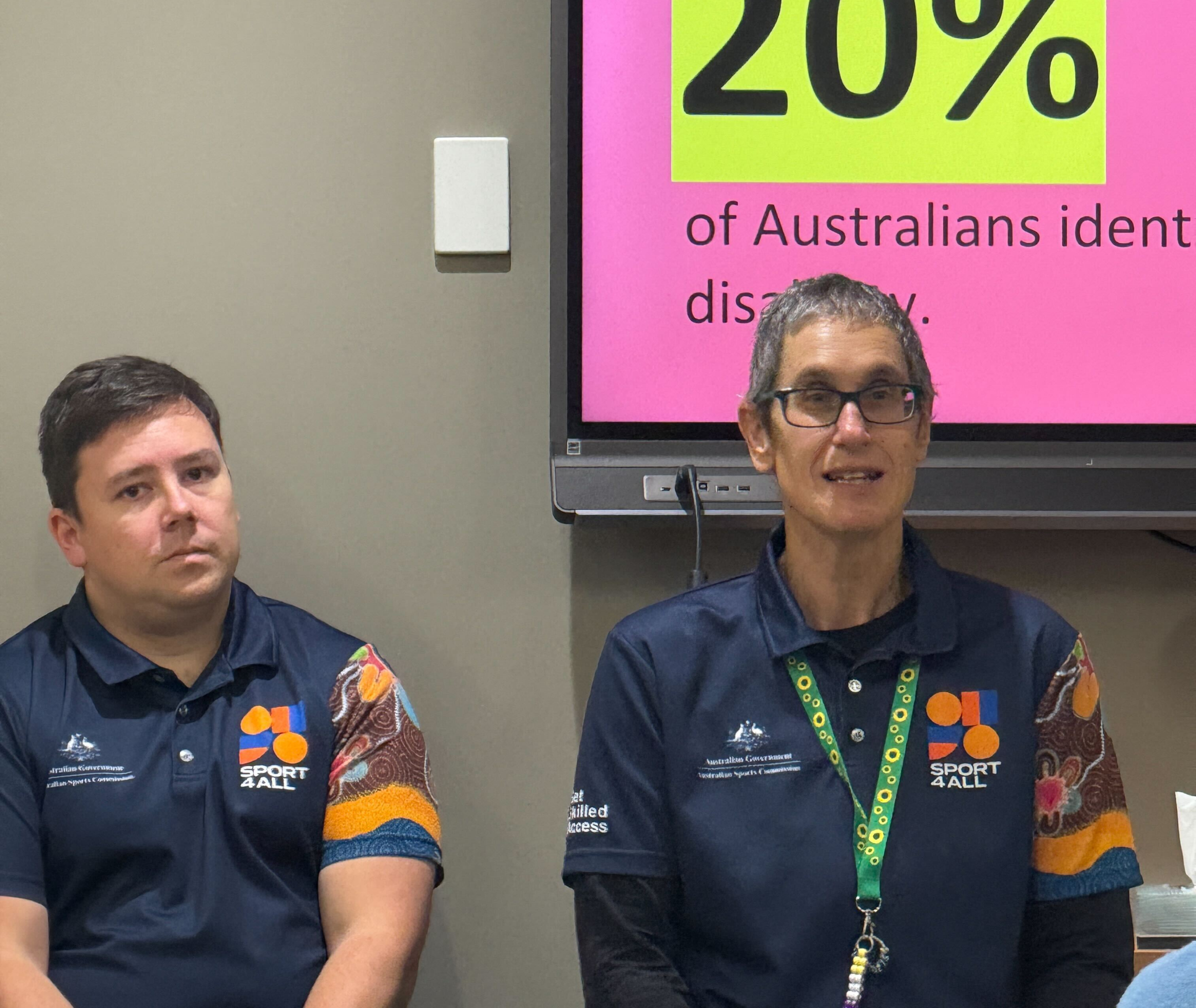 Two presenters in Sport4All uniforms seated in front of a bright pink slide displaying text about 20% of Australians identifying with disability.