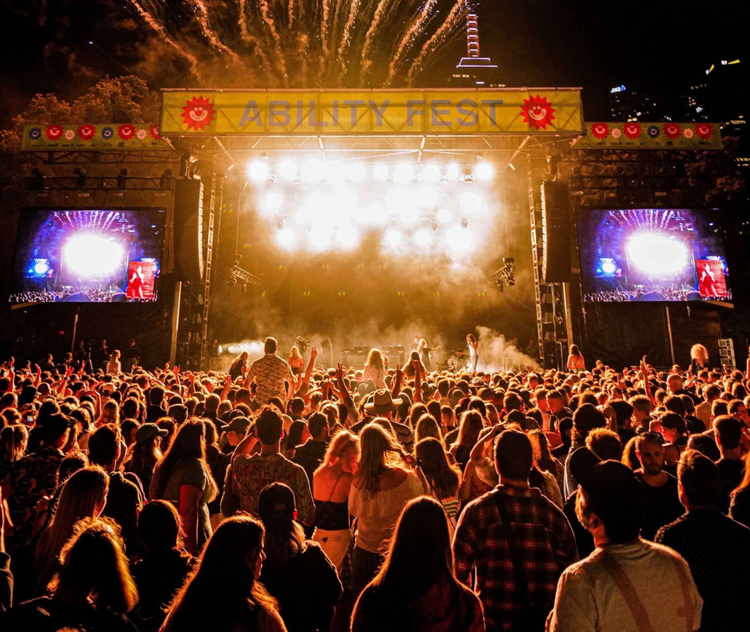 A large crowd at a live concert raises their hands toward a brightly lit stage.
