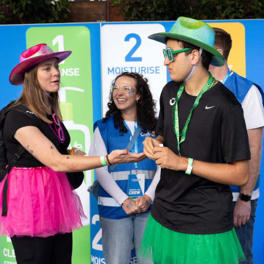 Three people in colorful outfits stand near a large bottle of CeraVe Hydrating Cleanser, with a banner in the background indicating skincare steps.