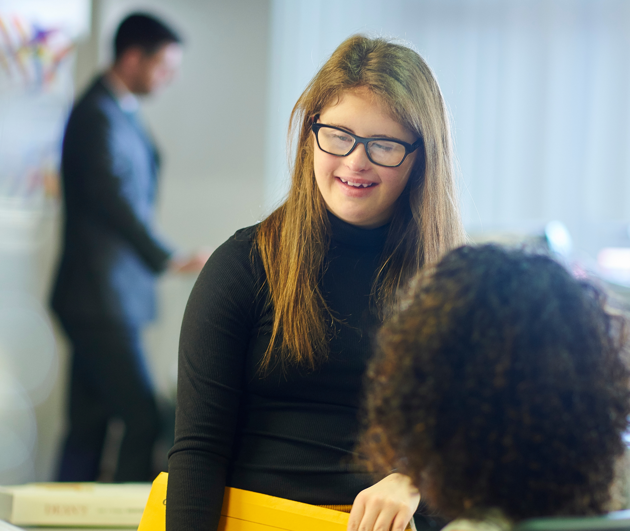 Young woman with long hair and glasses smiling while standing beside a bright yellow file folder in a office, with a blurred coworker in the background.